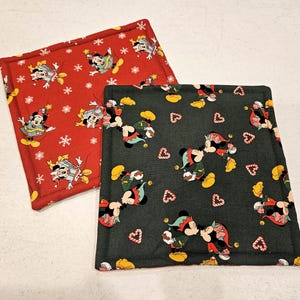 May include: Two fabric pot holders with a Christmas theme. One is red with white snowflakes and features Mickey Mouse and Minnie Mouse dressed as elves. The other is dark green with white hearts and features Mickey Mouse and Minnie Mouse kissing.