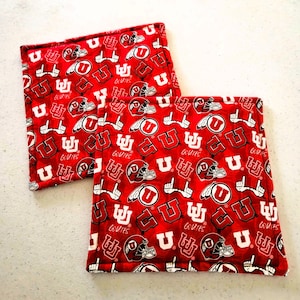 May include: Two red and white square pot holders with a repeating pattern of the letter "U", football helmets, and the words "GO UTES". The pot holders are made of a soft fabric.