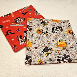 Puede incluir: Dos posavasos cuadrados de tela con temática de Disney. Uno es rojo con temática navideña y la palabra "CHEER" impresa en él. El otro es gris con temática de Halloween y presenta un patrón de telaraña.