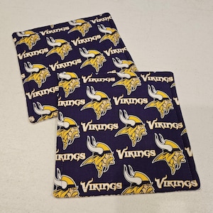 Puede incluir: Dos posavasos de tela morada con un patrón repetido del logotipo de los Minnesota Vikings. El logotipo es amarillo y blanco con un casco con cuernos.