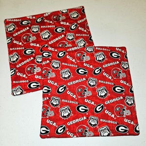May include: Two square red fabric coasters featuring a repeating pattern of the Georgia Bulldogs logo, helmets, and the words "GEORGIA", "UGA", and "BULLDOGS" in white and black. These coasters are likely for home use.