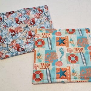 May include: Two blue and white fabric pot holders with a beach theme. One pot holder features a seashell, starfish, and seahorse design. The other pot holder features a surfboard, starfish, and seahorse design with the text "Fresh Fish" and "Beach".