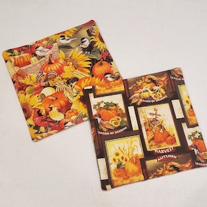 以下が含まれることがあります： 秋の収穫をテーマにした布製の鍋つかみ2枚。1枚は、かぼちゃ、かぼちゃ、ひまわりが入った豊穣の角。もう1枚は、かぼちゃ、ひまわり、そして「分かち合いの季節」、「秋の収穫」、「リンゴ」の文字が書かれたパッチワークのデザインです。