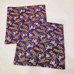 May include: Two square fabric coasters featuring a purple background with a repeating pattern of yellow text reading ECU PIRATES and EAST CAROLINA, interspersed with white and black skull and crossbones pirate logos wearing a purple hat.