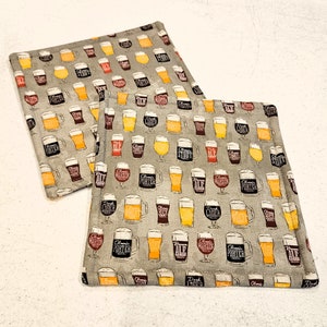 May include: Two beige fabric pot holders with a pattern of colorful beer mugs. The mugs have different beer styles printed on them, including "Classic Ale", "Dark Lager", "Stout", "Porter", and "Pale Ale".