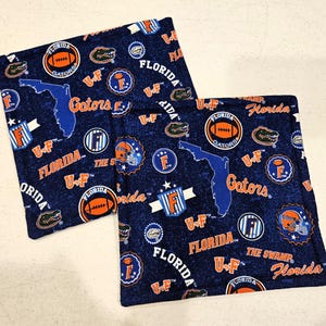 May include: Two blue fabric pot holders with a University of Florida Gators theme. The pot holders feature a repeating pattern of the Gators logo, the state of Florida, and the words "Florida", "Gators", and "U-F".