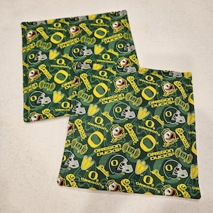 May include: Two square fabric coasters featuring a repeating pattern of the Oregon Ducks logo, duck heads, football helmets, and the text Oregon Ducks in yellow and white on a dark green background with stitched edges on all four sides.