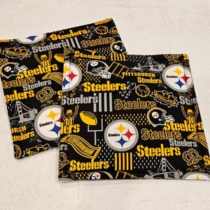May include: Two black and yellow fabric coasters with the Pittsburgh Steelers logo and team name repeated throughout. The coasters feature a variety of images including footballs, helmets, and the team's mascot.