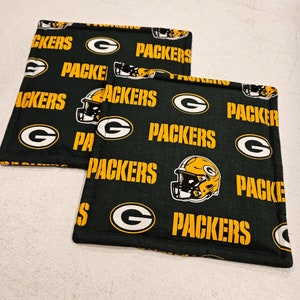 Op de afbeelding: Twee groene en gouden stoffen ovenwanten met de woorden "Packers" en "Packers" die erop worden herhaald. De ovenwanten hebben ook een groene "G" en een afbeelding van een voetbalhelm.