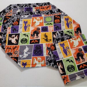 May include: Set of four Halloween-themed placemats with a black, orange, green, purple, and white patchwork design featuring ghosts, cats, pumpkins, bats, and witches. The placemats are made of fabric and have a stitched edge.