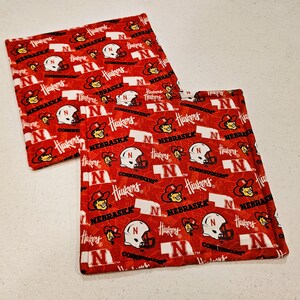 May include: Two red and white Nebraska Cornhuskers themed pot holders. The pot holders feature the team's logo, helmet, and the word "Nebraska" repeated across the surface.