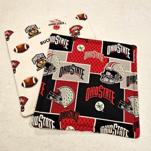May include: Two square fabric coasters. One is white with footballs and Ohio State logos. The other is red, black, and gray with Ohio State helmets and logos. The coasters are likely for drinks.
