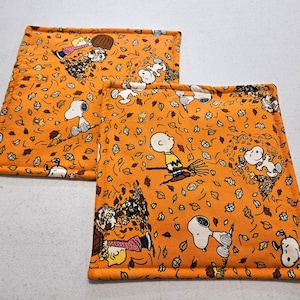 Op de afbeelding: Twee vierkante stoffen pannenlappen met een oranje achtergrond en een herhalend patroon van Snoopy, Charlie Brown en Woodstock die in herfstbladeren spelen. De doorgestikte items hebben gestikte randen en een zachte, gewatteerde constructie.