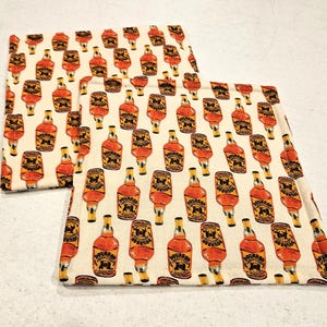 May include: Two white fabric coasters with a repeating pattern of orange and black bourbon bottles. The coasters are made of a soft, absorbent fabric and are perfect for protecting surfaces from spills and scratches.