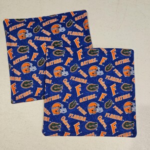 May include: Two blue fabric squares with a repeating pattern of orange football helmets, alligators, and the word "Gators" in white and orange. The word "Florida" is also visible. The squares are likely part of a set, possibly for kitchen or home decor.