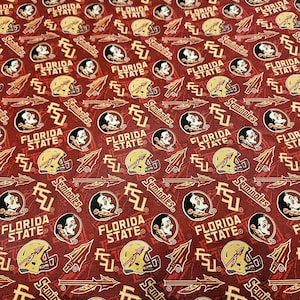 May include: A maroon fabric featuring a repeating pattern of Florida State Seminoles logos, including the team mascot profile, gold football helmets, the FSU monogram, the word Seminoles, and spear icons printed in gold, white, and black colours.