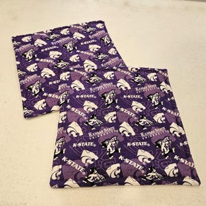 May include: Two square fabric pot holders featuring a purple background with a repeating pattern of the Kansas State University logo, the text K-STATE, and a mascot character holding a flag. The quilted items have stitched edges.