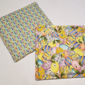 Könnte beinhalten: Zwei gequiltete Topflappen, einer mit hellblauem Hintergrund und einem Muster aus bunten Gummibärchen, der andere mit gelbem Hintergrund und einem Muster aus bunten Ostereiern.