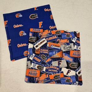 May include: Two square fabric items with a blue background. One has a pattern of orange football helmets, the letter "F", and the word "Gators". The other has a pattern of Florida license plates with the words "Gators", "Florida", and "Pride".