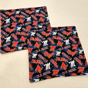 May include: Two square fabric items with a repeating pattern of "Ole Miss" and "Rebels" text, along with the letter "M". The text and "M" are in red and white on a navy blue background.
