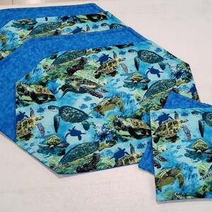 Op de afbeelding: Set van vier blauwe en groene achtkantige placemats met een ontwerp van zeeschildpadden en vissen. Twee bijpassende blauwe en groene vierkante onderzetters met hetzelfde ontwerp.