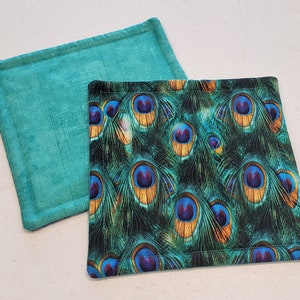 Puede incluir: Dos agarraderas de olla de tela. Una es verde turquesa y la otra tiene un patrón de plumas de pavo real.