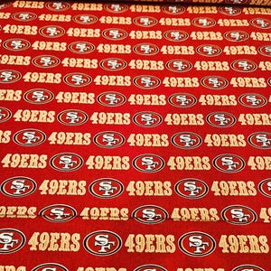 Peut inclure: Tissu rouge avec un motif répété du logo des 49ers de San Francisco et le texte "49ERS" en or. Idéal pour divers projets de couture et de bricolage.
