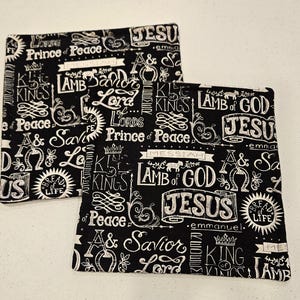 Puede incluir: Dos cuadrados de tela negros con texto blanco con frases religiosas como "Cordero de Dios" y "Jesús". Los cuadrados tienen un patrón repetido de palabras y elementos decorativos.