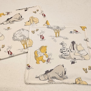 May include: Two square fabric items with a cream background featuring a Winnie the Pooh design. The illustrations include Pooh, Piglet, Eeyore, Tigger, and other characters. The edges are finished with a white border.
