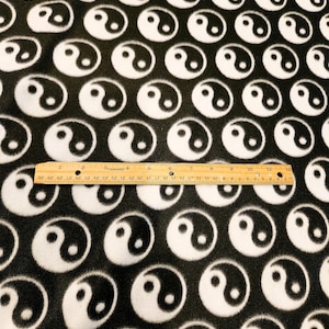 May include: Black fabric with a repeating pattern of white yin yang symbols. A wooden ruler, approximately 12 inches long, is placed across the fabric. The fabric has a soft, textured appearance.