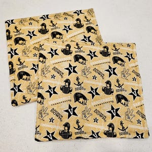 May include: Two square beige fabric items with a repeating pattern of black stars, anchors, and the text "Vanderbilt Commodores" and "Anchor Down". The fabric also features images of a person in a hat and a hand making a peace sign.
