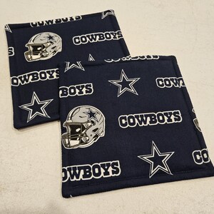 以下が含まれることがあります： 白い星と「COWBOYS」という文字が繰り返し書かれた、紺色の布製の鍋つかみが2つ。