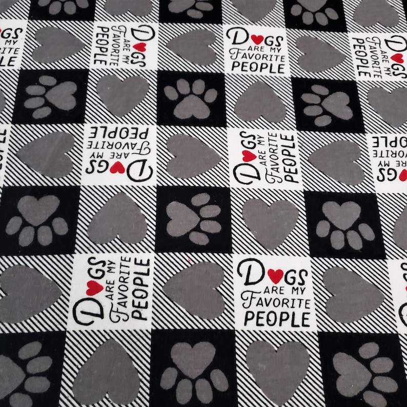 Op de afbeelding: Zwart-wit geblokt stof met een herhalend patroon van poot afdrukken, harten en de tekst "Honden zijn mijn favoriete mensen".