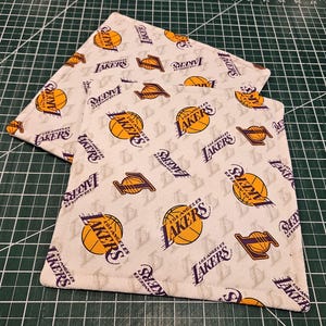 May include: Two square fabric coasters with a repeating pattern of the Los Angeles Lakers logo in gold and purple on a cream background. The coasters are made of a soft, likely absorbent material.