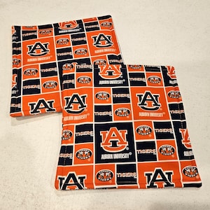 May include: Two square fabric pot holders featuring a repeating pattern of the Auburn University logo, the word Tigers, and the Auburn University text in navy blue and orange. The design consists of alternating navy blue and orange blocks.