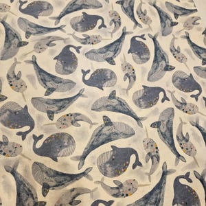 May include: A fabric featuring a repeating pattern of whales and narwhals in shades of blue and gray on a cream background. The whales have striped details and small pink accents. The design is whimsical and suitable for various craft projects.
