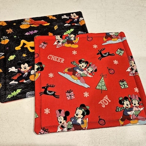 May include: Two red and black fabric placemats with a Christmas theme featuring Mickey Mouse and Minnie Mouse. The placemats have a white snowflake pattern and the words "Cheer" and "Joy" printed on them.