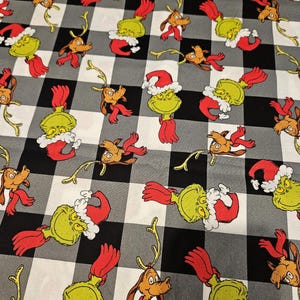 May include: Fabric with a black and white checkered pattern featuring cartoon characters. The characters include a green Grinch wearing a Santa hat and a brown dog with antlers and a red scarf. The fabric is likely for holiday-themed crafts.