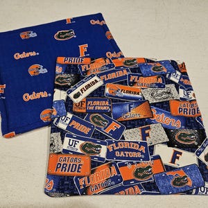 May include: Two blue fabric items with orange and white designs. One features the word "Gators" and helmet graphics. The other has license plate graphics with "Florida Gators" and "Pride" text. Both items are likely related to the University of Florida.
