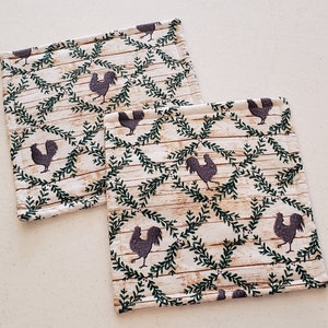 May include: Two cream-coloured fabric pot holders with a repeating pattern of green leafy branches and black roosters.