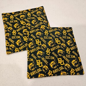 May include: Two square fabric coasters featuring a repeating pattern of the Baylor University logo, a bear mascot head, the word Baylor, and the phrase Sic 'Em in yellow ink on a black background. The coasters are flat with finished stitched edges.