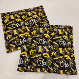 May include: Two square fabric items with a repeating pattern of the word "IOWA", the Hawkeyes logo, and football helmets in black, yellow, and white on a gray background. The items are likely coasters or small decorative squares.