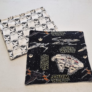 May include: Two black and white fabric pot holders. One features a repeating pattern of Stormtroopers from Star Wars. The other features a black background with stars and spaceships from Star Wars. The text "Star Wars" is printed on both pot holders.