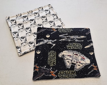 Könnte beinhalten: Zwei schwarze und weiße Stoff-Topflappen. Einer zeigt ein sich wiederholendes Muster von Stormtroopers aus Star Wars. Der andere zeigt einen schwarzen Hintergrund mit Sternen und Raumschiffen aus Star Wars. Der Text "Star Wars" ist auf beiden Topflappen gedruckt.