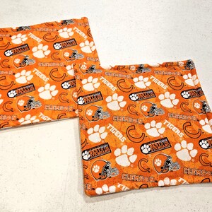 May include: Two square fabric pot holders featuring an orange background with a repeating pattern of white paw prints, orange football helmets, the letter C, the word Tigers, and the text Clemson Best is the Standard in black and white.
