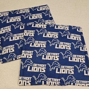 Pode incluir: Dois quadrados de tecido azuis com um padrão repetido das palavras "DETROIT LIONS" e um logotipo de leão branco. Os quadrados são provavelmente parte de um conjunto, possivelmente para decoração de casa ou artigos desportivos.