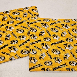 May include: Two yellow fabric squares with a repeating pattern of black and gold Missouri Tigers logos and text. The text includes "MISSOURI", "MIZZOU", and "TIGERS".