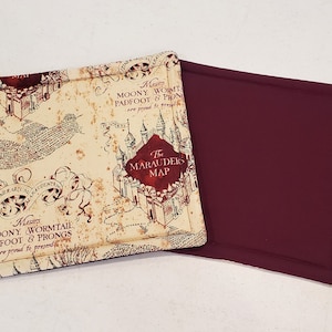 May include: A brown and beige fabric coaster with a map design featuring the text "The Marauders Map". The back of the coaster is a solid burgundy color.