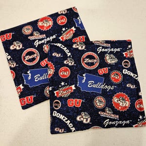 May include: Two navy blue Gonzaga University themed fabric squares. The squares are covered in Gonzaga logos, the word "Zags", and the word "Gonzaga" in white script. The state of Washington is also featured in blue.