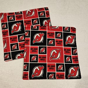 Könnte beinhalten: Zwei quadratische Stoffartikel mit einem sich wiederholenden Muster des New Jersey Devils-Logos in Rot, Schwarz und Weiß. Das Logo zeigt ein stilisiertes "N" und einen Hockeypuck. Die Worte "NEW JERSEY DEVILS" sind ebenfalls sichtbar.